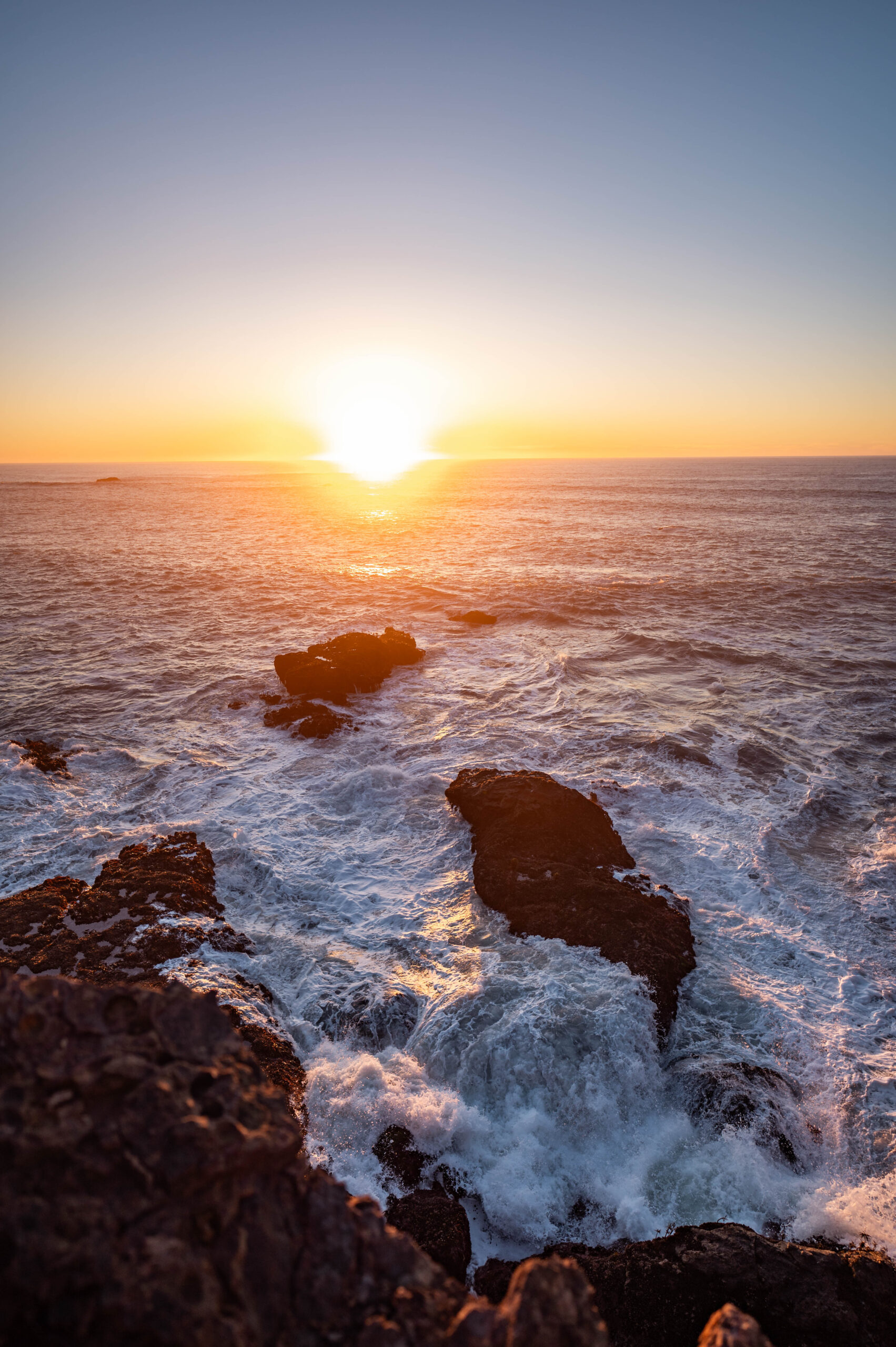 Waves of the Pacific Ocean crashing on the cliffside rocks in Northern California as the sun sets, creating a very orange and glowy image